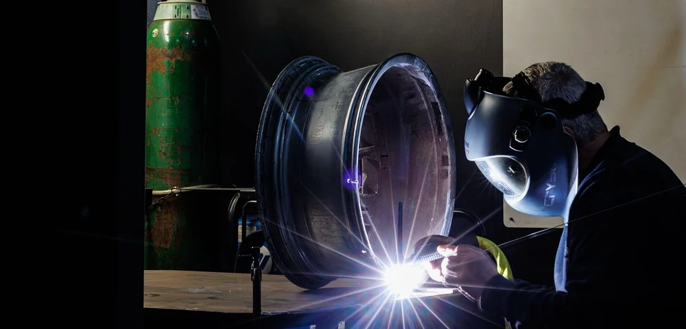 A technician welds a metal wheel rim while wearing a protective welding mask in a workshop.